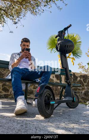 Young man looks at his smartphone sitting on a park bench next to his electric scooter Stock Photo