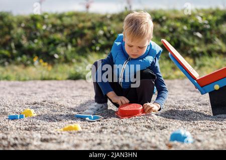 A little boy with a blue vest plays outdoors with small pebbles and colorful toys. Leisure time in the fresh air, a healthy child concentrates on play Stock Photo