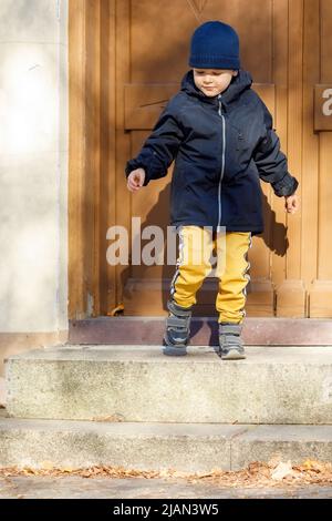 The little boy opens the big brown entrance door. The child leaves the house for an autumn walk. Stock Photo