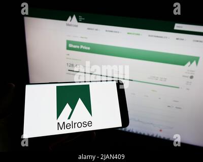 Person holding mobile phone with logo of British company Melrose Industries plc on screen in front of business web page. Focus on phone display. Stock Photo