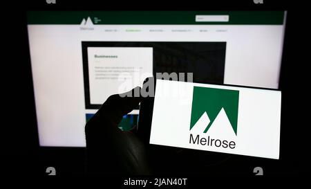 Person holding cellphone with logo of British company Melrose Industries plc on screen in front of business webpage. Focus on phone display. Stock Photo