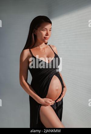 Asian pregnant woman model posing in black dress showing pregnancy bump in third trimester wearing jewelry. Vertical portrait in studio