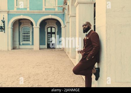 A dapper handsome African American bald bearded guy in a dark-red tailored costume is leaning against the wall, looking away and holding eyeglasses in Stock Photo