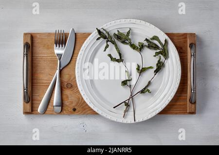 White ceramic plate and dry mint leaves and a tray with cutlery on a light painted wooden table. Mockup for demonstrating a dish Stock Photo