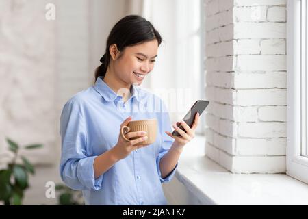 Positive asian woman drinking coffee and using smartphone Stock Photo