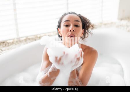 Pretty millennial woman blowing bubbles in foamy bath, playing in hot water at home Stock Photo