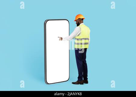 Black Builder In Hard Hat Touching White Screen On Huge Blank Smartphone Stock Photo