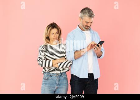 Offended Wife Looking At Husband As He Texting, Pink Background Stock Photo