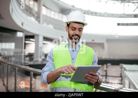 Cheerful young muslim engineer builder with beard in protective clothing typing on tablet, checking work Stock Photo
