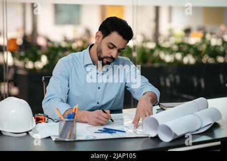 Serious smart busy young muslim man engineer architect with beard works with project drawings at workplace Stock Photo
