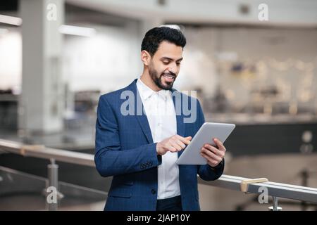 Cheerful confident young arab general manager with beard in suit works on tablet in office interior Stock Photo