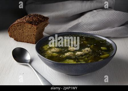 Rustic zucchini soup with meatballs with a piece of black bread with caraway seeds on a white table against the background of a gray kitchen towel in Stock Photo