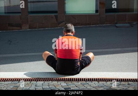 A lonely man in an orange-and-black cycling jersey sits on the floor in a sunny day Stock Photo