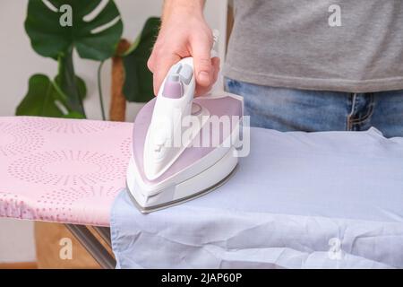 A man is ironing a shirt with an electric iron on an ironing board. Stock Photo