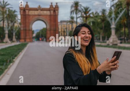 A fresh latin lady laughing while using his phone with an unfocusing arc of triomphe in the background Stock Photo
