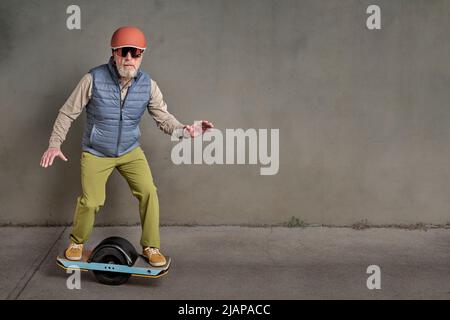 portrait of senior male hipster in helmet and goggles is riding one-wheeled electric skateboard in a grunge urban environment, copy space Stock Photo