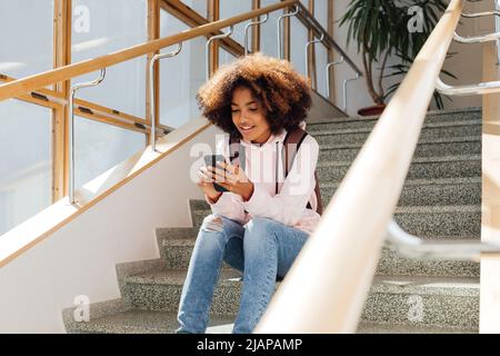 Smiling female student with backpack sitting on stairs and using mobile phone Stock Photo