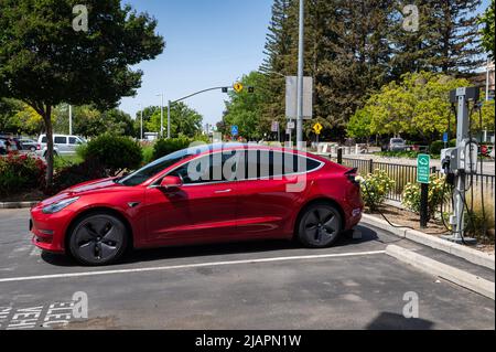 Photo of a red Tesla Model 3 with purple carpool lane sticker hooked up to an EV charger in a parking lot in East Sacramento. Stock Photo