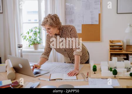 Senior woman architect with model of houses looking at blueprints in office. Stock Photo