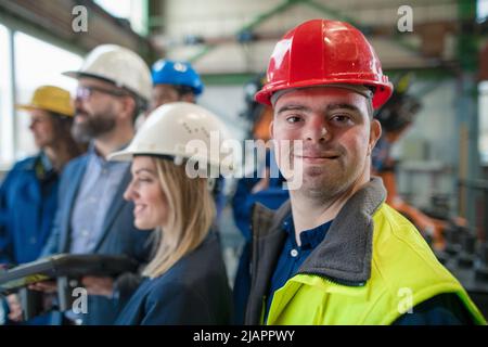 Young man worker with Down syndrome with manager and other collegues working in industrial factory, social integration concept. Stock Photo