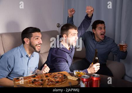 Happy men emotionally watching tv with beer and pizza at home Stock Photo