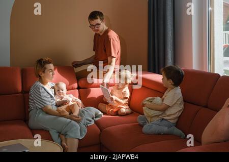 Big family on couch at home. Parents with kids on sofa during sunny day spending free time. Mother and father with three children reading book. Modern Stock Photo