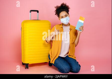 Full length photo of happy african american girl with medical mask on her face, holding passport and travel tickets, sitting near big yellow suitcase on isolated pink background, looks at camera Stock Photo
