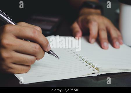 Man holding a pen and writing on a notebook on his desk. Stock Photo