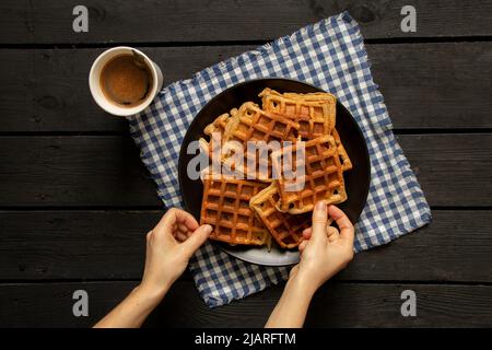 female hands take Viennese waffles on a wooden table and a cup with coffee, waffles for breakfast in the kitchen at home, there are waffles Stock Photo