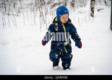 Joyful curious and independent toddler kid walks along the path in the winter forest. A little boy in warm overalls and a hat enjoys a walk in the win Stock Photo