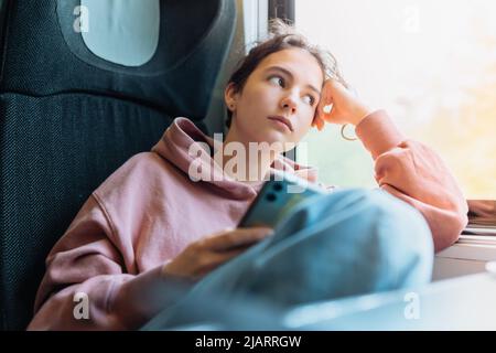 pensive lonely teenage girl sits in train near window, resting head on hand, looking through glass, holding smartphone in hands, bending knees Stock Photo
