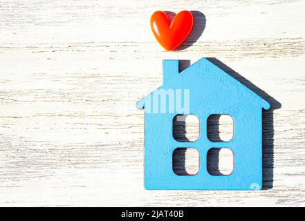 Flat plywood house shape painted blue with a small red heart on a wooden background with copy space. Stock Photo