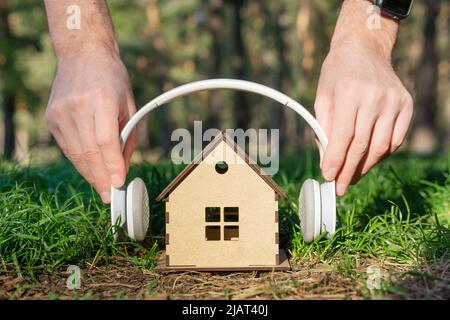 Crop view of male hands placing white wireless headphones on a miniature plywood house model in the forests. Creative home insulation concept. Stock Photo