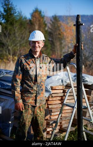 Male worker building pile foundation for wooden frame house. Portrait of man builder in white safety helmet drilling piles into the ground on sunny day. Stock Photo