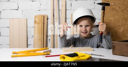 smiling little girl with helmet learn to hammer a nail in wooden plank at woodworking workshop. copy space Stock Photo