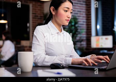 Confident asian businesswoman sitting in office workspace at night while working on laptop. Young adult entrepreneur researching startup business ideas while developing marketing strategy for project. Stock Photo
