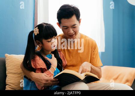 Asian dad embracing daughter and reading fairytale book while sitting on sofa near window at home Stock Photo