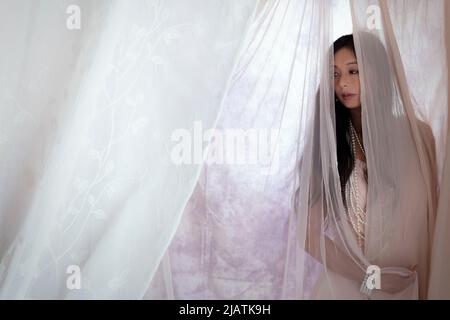 A beautiful Asian woman wearing a pink vintage dress and posing amongst draped sheer fabric Stock Photo