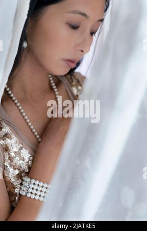 A beautiful Asian woman wearing a gold sequin top and tulle skirt and posing amongst draped fabric Stock Photo