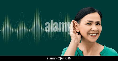 Positive woman with hand near ear and with luminous sound wave on green background showing variety of sounds going to her ear. Auditory health concept Stock Photo
