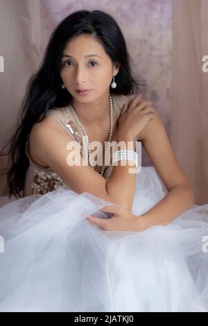 A beautiful Asian woman wearing a gold sequin top and tulle skirt and posing amongst draped fabric Stock Photo