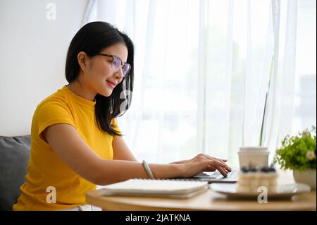 Attractive asian millennial woman with an eyeglasses sits in the modern bright workspace, woking on her project, using laptop computer. Stock Photo