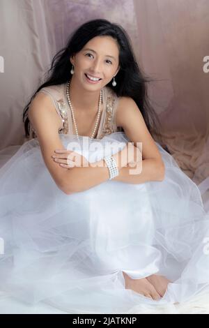 A beautiful Asian woman wearing a gold sequin top and tulle skirt and posing amongst draped fabric Stock Photo