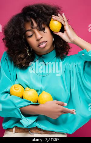 brunette african american woman in turquoise blouse posing with lemons isolated on pink Stock Photo