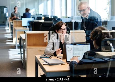 Colleagues cooperating with each other behind glass window at office, they discussing graphs on computer Stock Photo