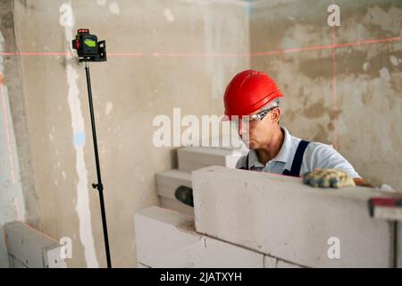 Bricklayer using rubber mallet hammer to tap and level concrete blocks wall. Contractor uses precise spirit bubble and laser level for brickwork Stock Photo
