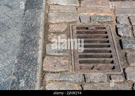 rusted rectangular grating manhole drainage system on the pavement road nearby to the gray granite curb and stone tile pedestrian pavement close-up on Stock Photo