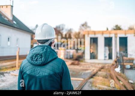 40s bearded engineer worker with helmet on head standing outside of building in construction process, taking photo on smart phone  Stock Photo