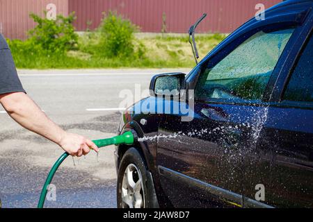 Man is washing car outdoor. Male hand holds hose with water and waters vehicle. Cleaning car in garage in front of house. Stock Photo