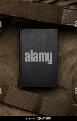 A closed book with a black cover on an old hiking backpack. Vintage khaki canvas background. Stock Photo